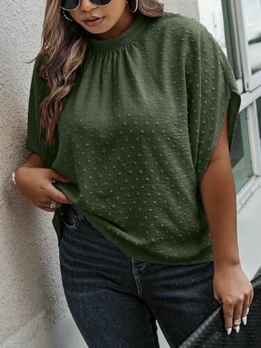 Woman in a green top and jeans, standing against a wall.